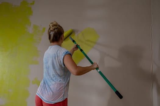 A woman is diligently using a roller to apply bright yellow paint onto a wall in the interior of her home