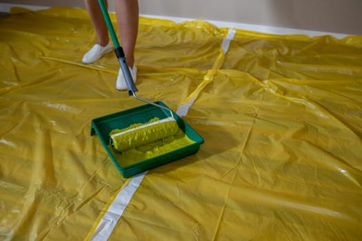 A person is meticulously painting a wall with a roller, while using a yellow drop cloth to ensure protection