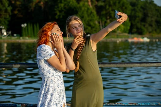 Two friends joyfully take a selfie by the serene water on a beautiful sunny day, capturing special moments together