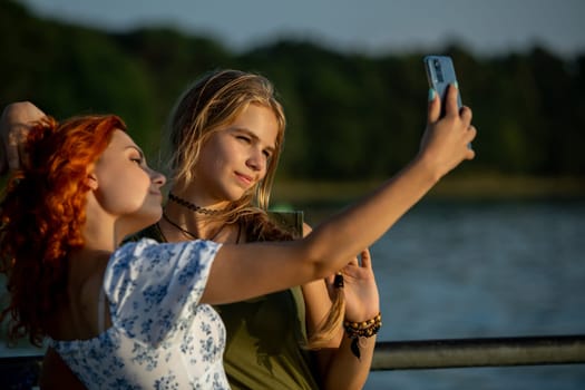 Two young women joyfully capturing a fun and lively selfie by the beautiful water during golden sunlight hours