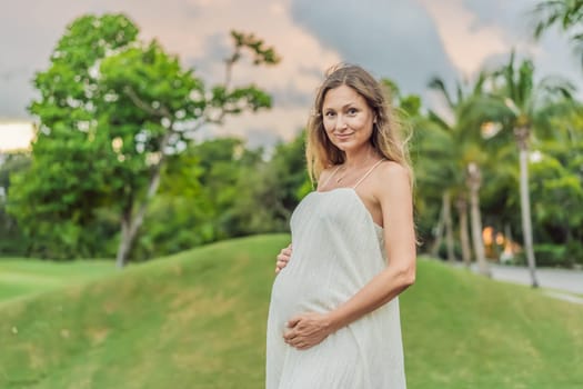 Pregnant woman hugging her tummy standing outdoors surrounded by nature. Pregnancy, expectation, motherhood concept.