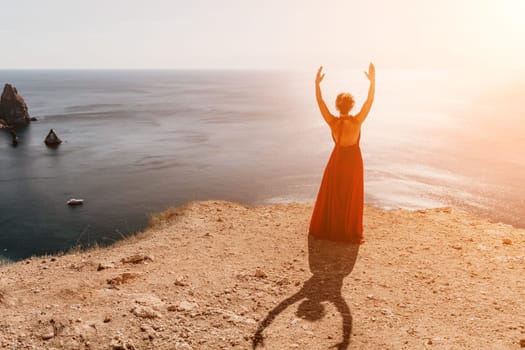 Side view a Young beautiful sensual woman in a red long dress posing on a rock high above the sea during sunrise. Girl on the nature on blue sky background. Fashion photo.