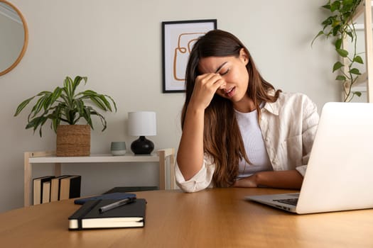Young woman feeling exhausted and overworked using laptop in the office. Headache and burnout concept.