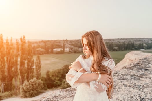 Romantic beautiful bride in white dress posing with sea and mountains in background. Stylish bride standing back on beautiful landscape of sea and mountains on sunset