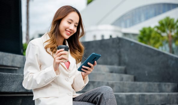 Lifestyle woman business people using mobile smart phones in the city outdoors office building. Young Asian female wearing blue shirt while use cellphone with holding coffee cup sitting on staircase