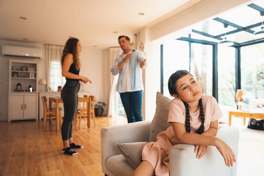 Annoyed and unhappy young girl sitting on sofa trapped in middle of tension by her parent argument in living room. Unhealthy domestic lifestyle and traumatic childhood develop to depression.Synchronos