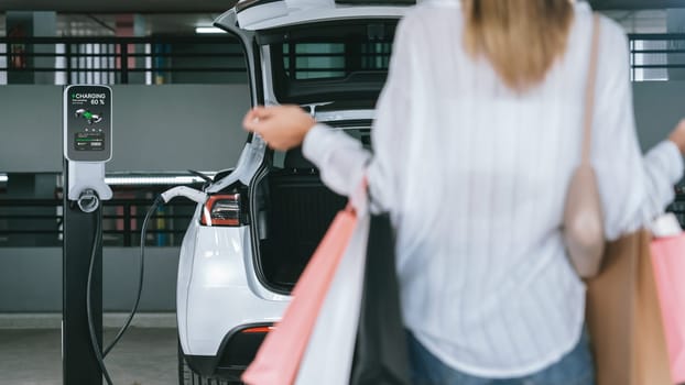 Young woman travel with EV electric car to shopping center parking lot charging in downtown city showing urban sustainability lifestyle by green clean rechargeable energy of electric vehicle innards