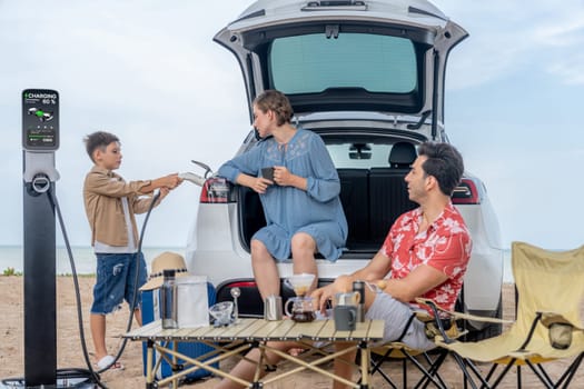 Family vacation trip traveling by the beach with electric car, happy family recharge EV car, enjoying outdoor camping coffee. Seascape travel and eco-friendly car for clean environment. Perpetual