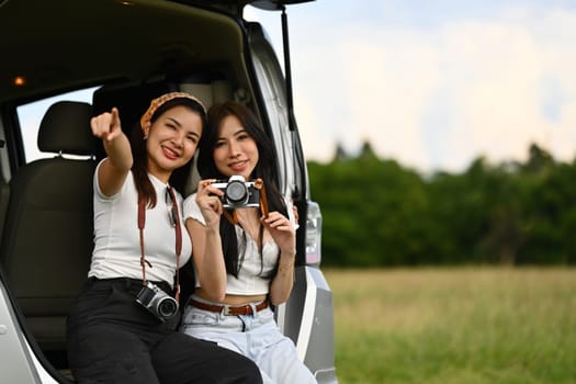 Two best girlfriends sitting in open car trunk and enjoying nature view. Road trip, traveling and lifestyle concept.