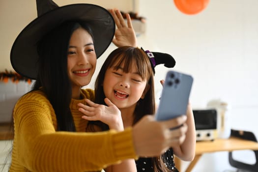 Smiling asian mother taking a selfie on smart phone and having fun with her daughter in Halloween decorated kitchen.