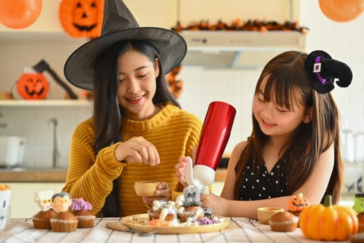 Smiling young mother and her little daughter getting ready for the holiday, making Halloween cupcakes.