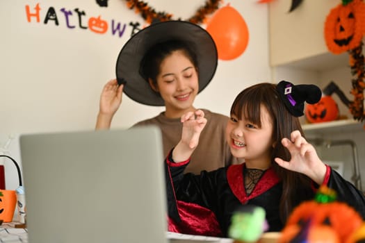 Cute little girl and mother dressed as a witch having online video call on laptop at home decorated for Halloween.