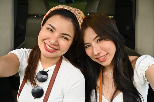 Happy best female friends taking a selfie while sitting on opened trunk of car. Journey, traveling and lifestyle concept.