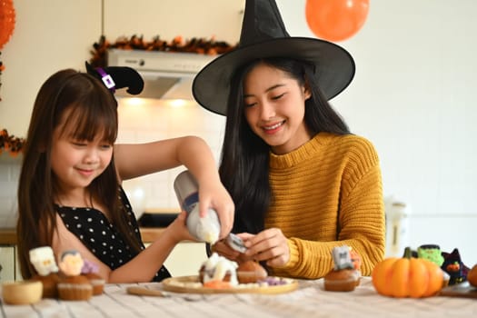 Happy mother with little daughter preparing delicious sweet dishes for Halloween party in kitchen.