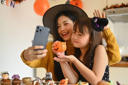 Happy young mother and her daughter taking a selfie on smart phone in Halloween decorated kitchen.