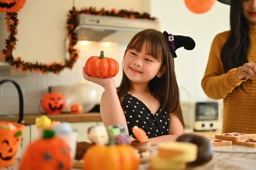 Adorable little Asian girl holding small pumpkin up by her face while preparing for holiday party with her mother in kitchen.