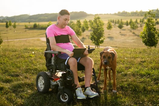 A young gay with a disability is seen enjoying time with his dog while using a tablet at the park at sunset.