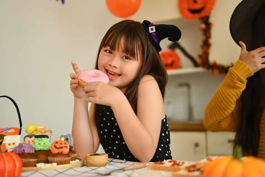 Cute little girl eating donut in Halloween decorated kitchen. Halloween celebration concept.