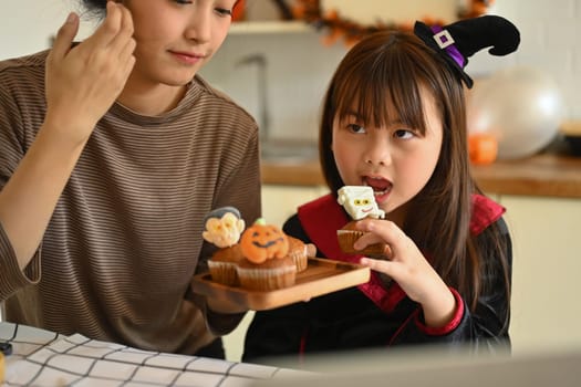 Cute little asian girl dressed as a witch eating colorful Halloween cupcakes with mother in kitchen. People, holiday and festival.