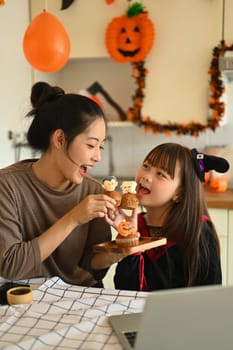 Cute little asian girl dressed as a witch eating colorful Halloween cupcakes with mother in kitchen. People, holiday and festival.