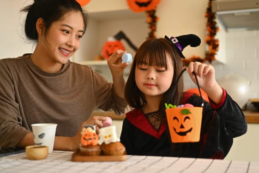 Smiling young mother and her little daughter getting ready for the holiday, making Halloween cupcakes.