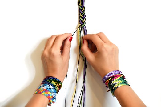 Female hands weaving knot for DIY friendship bracelet Pigtail. Many handmade bracelets on girls wrists. Friendship bracelet making process, step by step. White background with copy space
