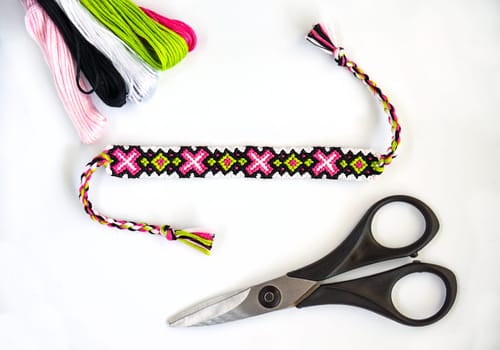 Woven handmade DIY friendship bracelets with bright pink and green pattern next to skeins of thread and scissors on white background