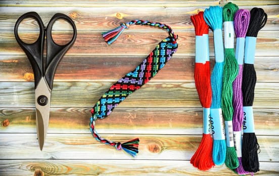 Woven DIY friendship bracelet with abstract pattern near skeins of embroidery floss, scissors on wooden background.