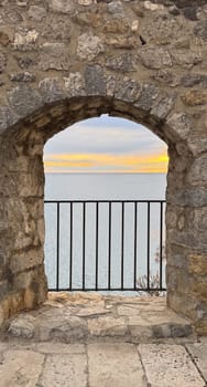 A rustic stone archway frames a view of calm blue waters and a golden sunset, with a black iron railing in the foreground. High quality photo