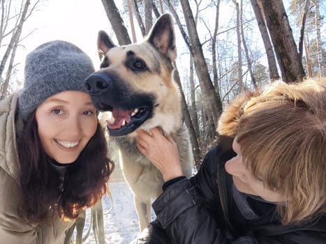Two women smile while interacting with a large dog in a snowy forest on a bright winter day, surrounded by trees.