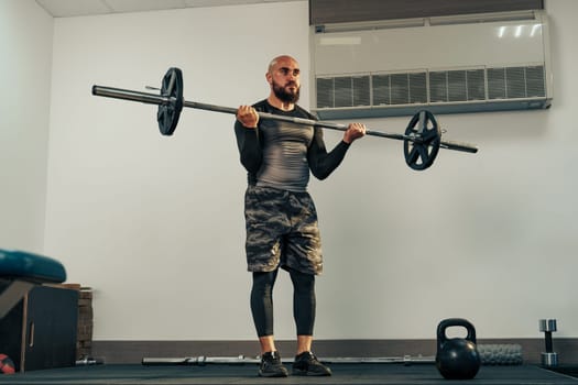 A man engaged in strength training holds a barbell at shoulder height in a gym. He focuses on his form while surrounded by gym equipment and weights.
