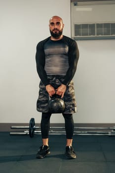 A fit man stands in a gym holding a kettlebell, wearing a fitted long-sleeve shirt and shorts. The gym features various exercise equipment and weights, indicating an active workout environment.