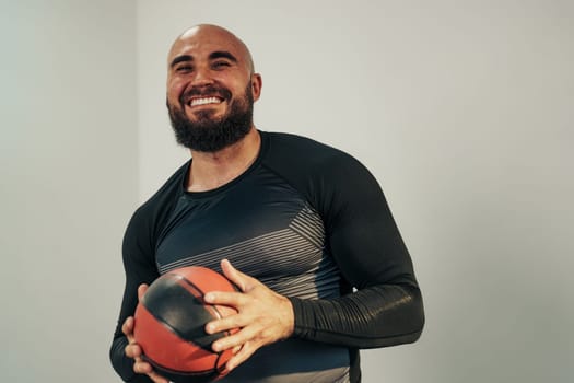 A man with a beard smiles while holding a basketball indoors. He wears a fitted black athletic shirt and stands against a plain wall, reflecting a positive energy during training.