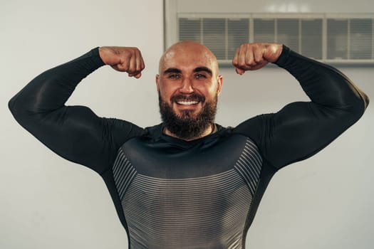 A muscular individual flexes his biceps with a confident smile in a gym environment. He wears a fitted athletic shirt, highlighting his physique and dedication to fitness.