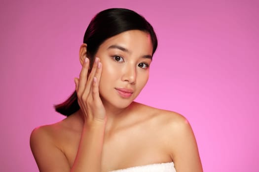 A woman gently touches her face while applying a skincare product. She has a serene expression and is illuminated by a vibrant pink background. The setting suggests a beauty routine.