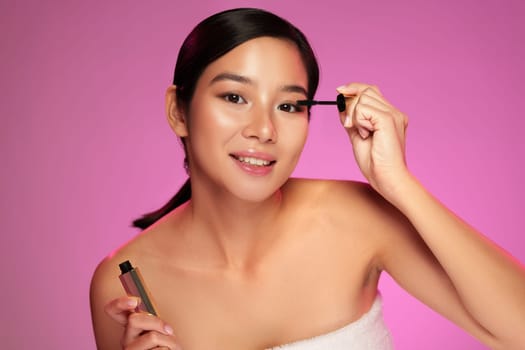 A model is applying mascara with a focused expression while holding a cosmetic tube. The background is vibrant pink, enhancing the beauty theme of the moment.