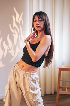A dancer showcases her skills in a bright room, wearing a black tank top and beige pants. Soft shadows from the light create an artistic ambiance as she strikes a pose.