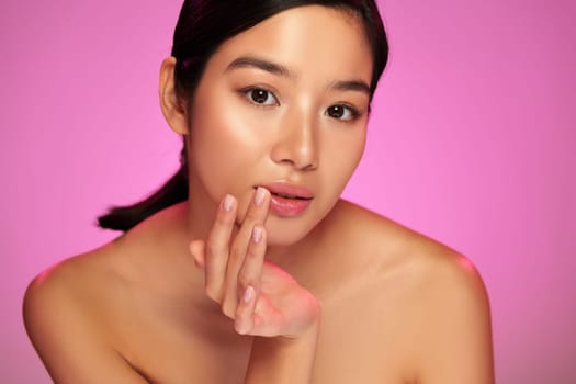 A model with a natural look poses against a bright pink backdrop. She softly touches her face, highlighting her smooth skin and graceful expression, embodying elegance and poise.