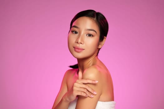 A young woman with smooth skin poses elegantly against a bright pink backdrop. She exudes confidence and tranquility while demonstrating a skincare routine with gentle gestures.