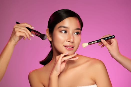 A model showcases a serene expression while receiving makeup application from two skilled artists. The backdrop is a bright pink, enhancing the beauty ritual atmosphere.