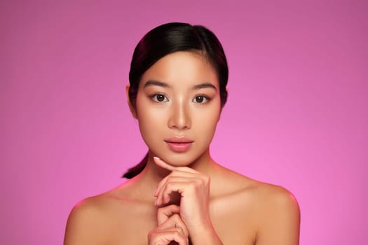 A model showcases a serene expression while resting her chin on her hands against a bright pink background. The studio lighting highlights her features, creating a soft and inviting look.