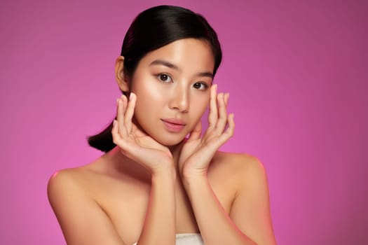 A young model poses elegantly, showcasing natural beauty with her hands gently framing her face. The vibrant pink background enhances her features, creating a striking visual contrast.