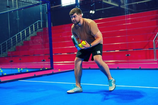 A male athlete stands poised with a paddle and ball, preparing to serve in an indoor padel match. The vibrant blue court contrasts with the red seating, creating an energetic atmosphere.