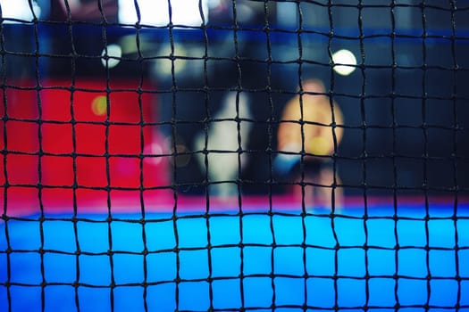 In a vibrant indoor sports facility, athletes train together against a backdrop of colorful surfaces. A net blurs the view of their activities, creating a dynamic atmosphere during the evening.