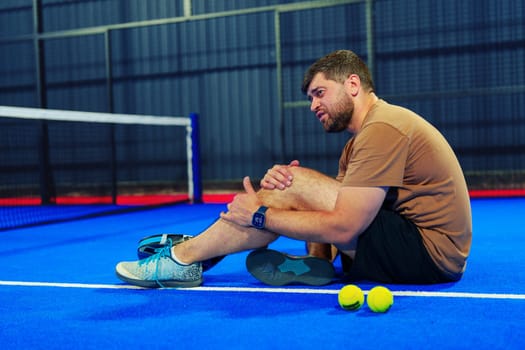 A man is seated on a vibrant blue tennis court, visibly in pain as he holds his ankle. Nearby, tennis balls lie on the ground, indicating a recent match.