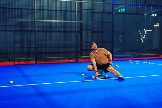 A man is engaged in an intense game of padel tennis, making a dynamic dive to reach a ball on a bright blue court. The atmosphere is lively and competitive as balls scatter around.