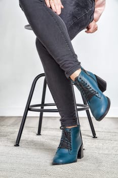 Model elegantly poses on a stool, highlighting fashionable teal ankle boots and stylish gray jeans in a contemporary environment.
