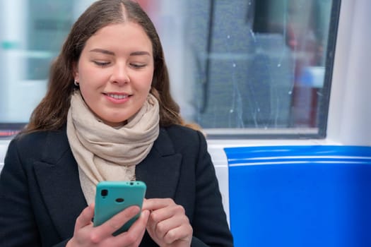 Cheerful young woman in a black coat and beige scarf sitting inside a modern subway train, laughing while looking at her smartphone. She enjoys a fun moment during her commute.
