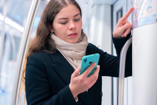 Focused young woman in a black coat and beige scarf checking a subway map while holding her smartphone. She navigates her route inside a modern metro train.