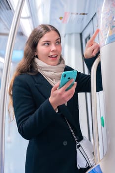 Focused young woman in a black coat and beige scarf checking a subway map while holding her smartphone. She navigates her route inside a modern metro train.